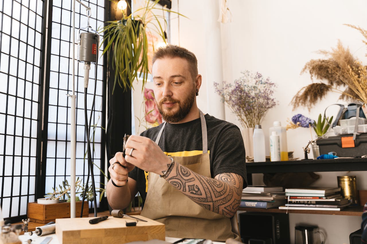 A skilled jewelry artisan working in a sunlit studio, surrounded by tools and greenery, wearing an apron.